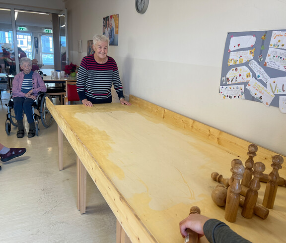 Bewohnerinnen spielen Tischkegeln an einer langen Holzbahn im Gemeinschaftsraum. 