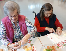 Zwei Seniorinnen sitzen am Tisch und schneiden Kartoffeln und Zwiebeln für das Essen.