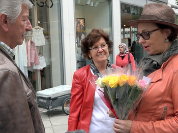 Drei Menschen treffen sich in der Stadt in Dornbirn. Eine Frau hält mehrere bunte Rosen in der Hand. Im Hintergrund sieht man ein Einkaufsgeschäft.