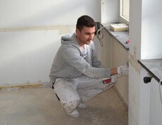 Ein junger Herr mit schwarzen kurzen Haaren, grauem Pullover und einer weißen Hose malt eine Wand weiß. Dabei steht er gebückt im Eck des Raums neben dem Fenster.
