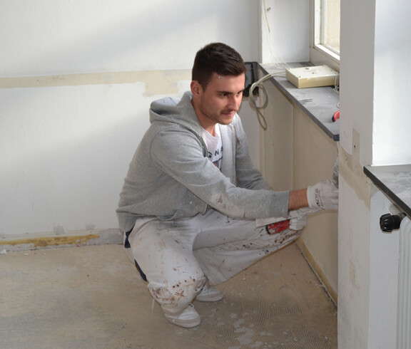 Ein junger Herr mit schwarzen kurzen Haaren, grauem Pullover und einer weißen Hose malt eine Wand weiß. Dabei steht er gebückt im Eck des Raums neben dem Fenster.