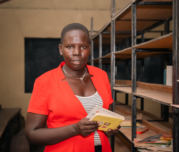 Eine Lehrerin mit orangem Jackett steht in einer Schulbibliothek und hält ein Buch in ihrer Hand.