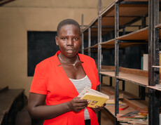 Eine Lehrerin mit orangem Jackett steht in einer Schulbibliothek und hält ein Buch in ihrer Hand.