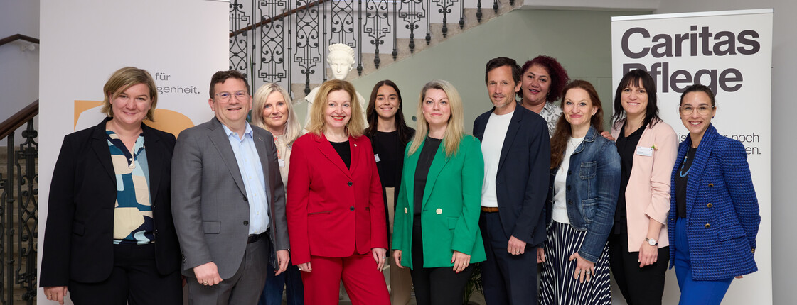 Gruppe von elf Personen steht für ein Gruppenfoto in einem Innenraum mit Treppe und Geländer; im Hintergrund sind zwei Roll-up-Banner zu sehen, eines mit der Aufschrift „Caritas Pflege“.