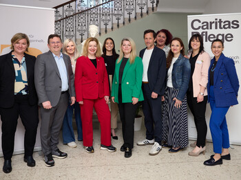 Gruppe von elf Personen steht für ein Gruppenfoto in einem Innenraum mit Treppe und Geländer; im Hintergrund sind zwei Roll-up-Banner zu sehen, eines mit der Aufschrift „Caritas Pflege“.