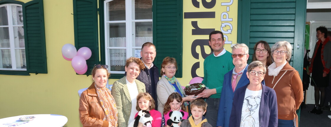 Eine Gruppe von Menschen steht vor einem gelben Gebäude mit grünen Fensterläden; einige Kinder halten Stofftiere, und in der Mitte wird eine dekorierte Torte präsentiert.
