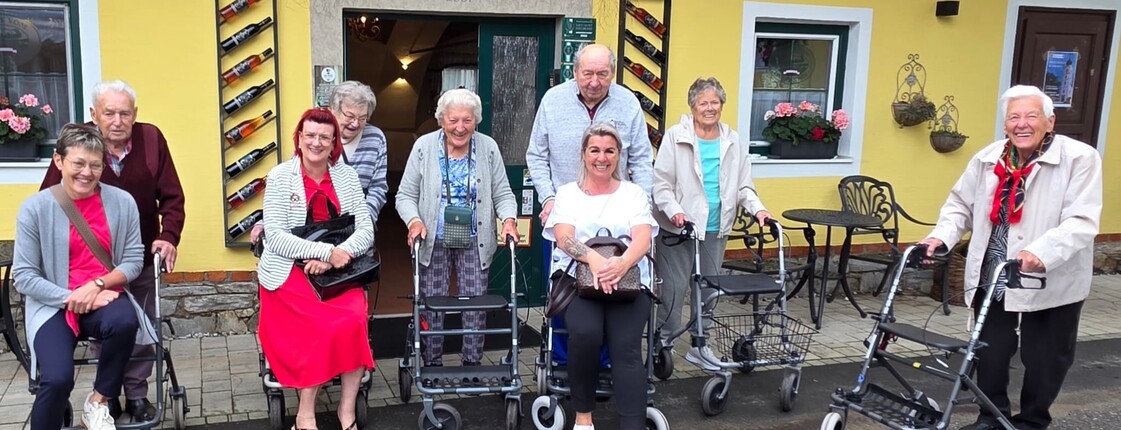 Bewohnerinnen und Bewohner eines Pflegewohnhauses haben gemeinsam einen Ausflug zu einem Weingut unternommen und stehen vor dem Gebäude für ein Gruppenfoto.