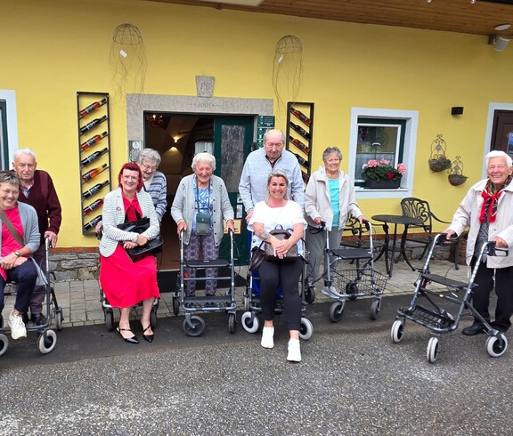 Bewohnerinnen und Bewohner eines Pflegewohnhauses haben gemeinsam einen Ausflug zu einem Weingut unternommen und stehen vor dem Gebäude für ein Gruppenfoto.