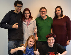Für ein Gruppenfoto stehen sechs junge Frauen und Männer zusammen. Zwei sind in der ersten Reihe kniend am Boden, die anderen vier stehen dahinter. Die beiden Männer tragen eine Brille. Gekleidet sind sie ganz unterschiedlich in verschieden Farben wie schwarz, beige, grün, rot und blau. Alle bis auf zwei machen für das Bild einen Daumen nach oben. 