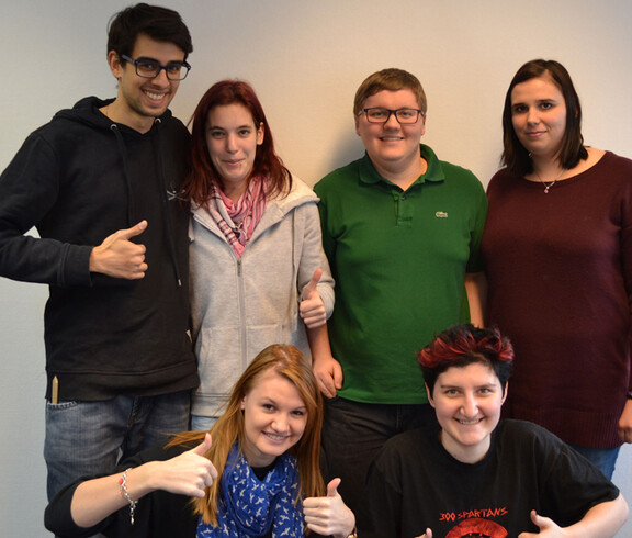 Für ein Gruppenfoto stehen sechs junge Frauen und Männer zusammen. Zwei sind in der ersten Reihe kniend am Boden, die anderen vier stehen dahinter. Die beiden Männer tragen eine Brille. Gekleidet sind sie ganz unterschiedlich in verschieden Farben wie schwarz, beige, grün, rot und blau. Alle bis auf zwei machen für das Bild einen Daumen nach oben. 