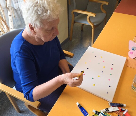 Eine weiter Bewohnerin des Pflegewohnhauses gestaltet konzentriert mit bunten Farbtupfern eine Leinwand und nimmt aktiv am kreativen Malangebot teil.