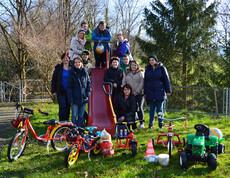Eine Gruppe junger Erwachsener steht auf einem Kinderspielplatz und zeigt stolz ihre Sachspenden für MUKI. Darunter sind unter anderem Fahrräder und Spielzeugtraktoren. 