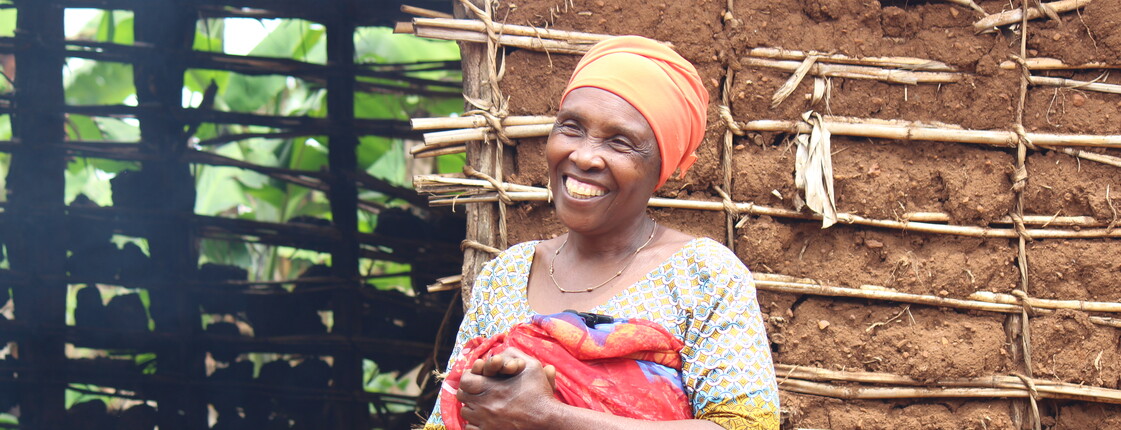 Portrait einer lachenden Frau in traditioneller Kleidung in Tansania, die vor einer Lehmhütte steht.