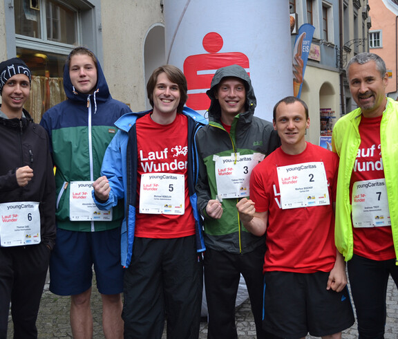 Sieben Caritas Mitarbeiter stehen zu einem Gruppenfoto in der feldkircher Innenstadt zusammen. Sie nehmen an einem Laufwettbewerb teil und haben dementsprechend überwiegend Sportbekleidung an. 