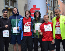 Sieben Caritas Mitarbeiter stehen zu einem Gruppenfoto in der feldkircher Innenstadt zusammen. Sie nehmen an einem Laufwettbewerb teil und haben dementsprechend überwiegend Sportbekleidung an. 