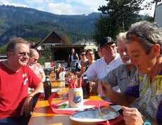 10 Personen sitzen an einem langen Biertisch und essen. Sie sind im Freien und haben es bei schönem Wetter gemütlich.  