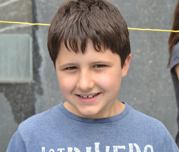 Ein Bub im Volksschulalter blickt in die Kamera. Er hat kurze schwarze Haare und trägt ein blaues Oberteil. Sein Gesicht ist ründlich. Er lächelt. 