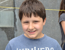 Ein Bub im Volksschulalter blickt in die Kamera. Er hat kurze schwarze Haare und trägt ein blaues Oberteil. Sein Gesicht ist ründlich. Er lächelt. 