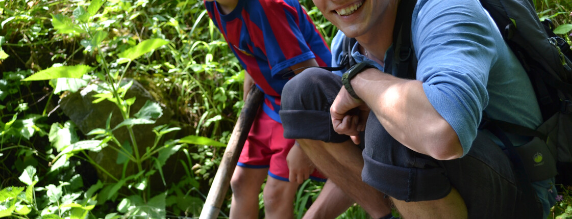Ein junger Erwachsener und ein Bub sind in der Natur. Der junge Bursche hat einen Fußballtrikot an und hält einen Stock in der Hand. Der Erwachsene trägt einen Rucksack und steht auf einem großen Stein. Beide haben kutze Haare. Der Hintergrund schücken grüne Gräser. 