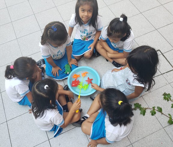 Sechs kleine Mädchen in blau-weißer Schuluniform sitzen im Kreis auf dem Boden und spielen gemeinsam mit bunten Spielzeugfischen in einer Schüssel mit Wasser.