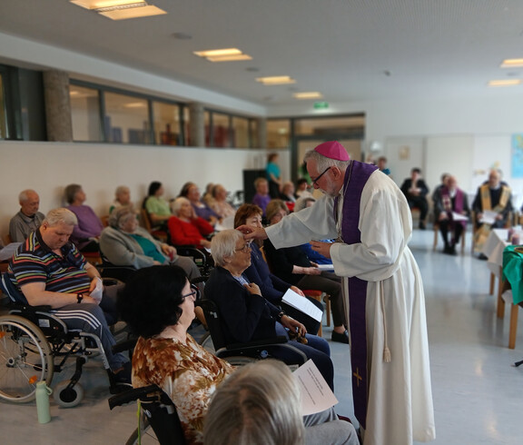Bischof Josef Marketz teilt das Aschenkreuz an sitzende Bewohner*innen im Saal des Haus Martha aus.