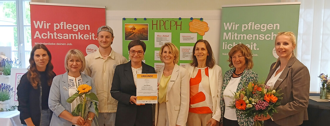 Gruppe aus Projektleitung, Pflege, Politik und Trägervertretungen steht mit Urkunde und Blumen vor HPCPH‑Tafel und Caritas‑Rollups