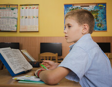 Ein kleiner junge sitzt an seinem Tisch in einem Klassenzimmer. Vor ihm steht ein offenes Lesebuch.