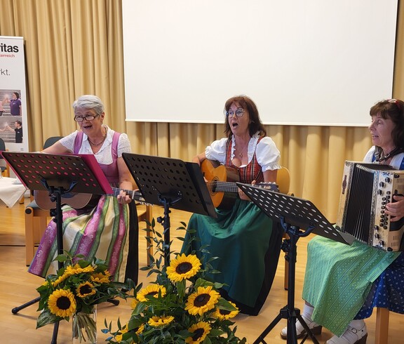Drei Frauen sitzen und spielen Musikinstrumente, zwei Gitarren und eine Harmonika.