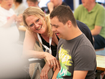 Eine Betreuerin beugt sich zu einem Menschen mit Beeinträchtigungen und lächelt ihn an.