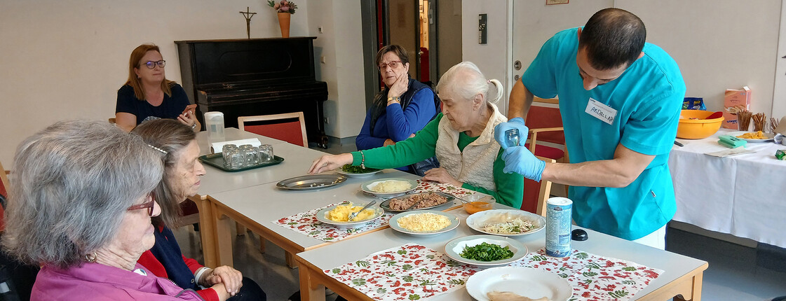 Ein Praktikant unterstützt eine Seniorin bei der Zubereitung eines tunesischen Gerichts am Tisch.