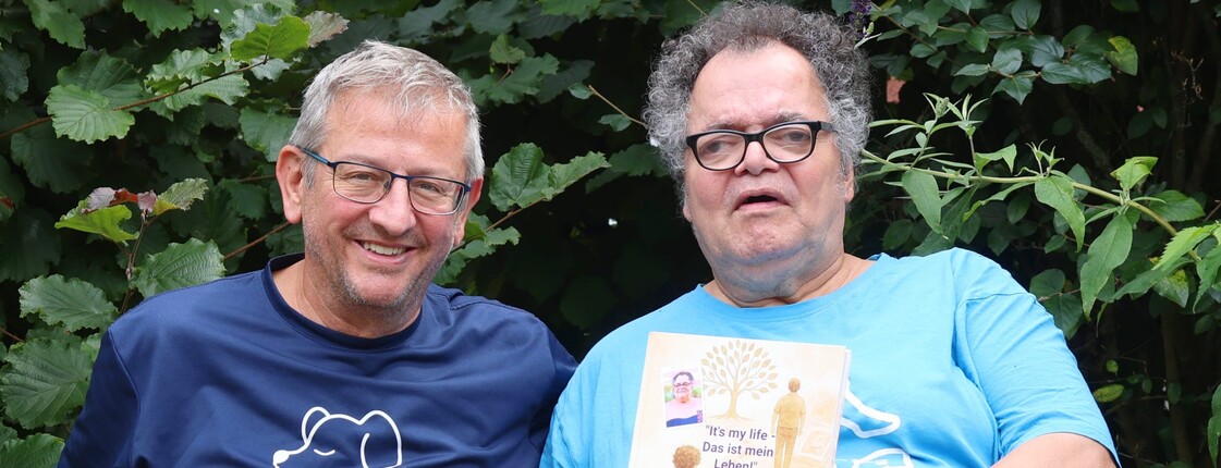 Zwei ältere Männer sitzen auf einer Bank und schauen in die Kamera. Der Mann mit Brille rechts hält ein Buch in der Hand.