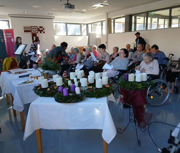 Mehrere Adventkränze mit Kerzen auf Tisch, Menschen sitzen dahinter und lesen