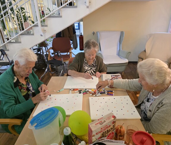 Drei Bewohnerinnen des Pflegewohnhauses sitzen gemeinsam am Tisch und gestalten mit bunten Farben kreative Punktbilder auf Leinwänden.