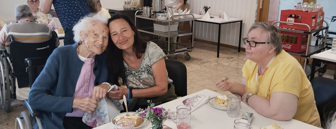 Eine Angehörige umarmt eine Bewohnerin liebevoll an einem gedeckten Tisch mit Torte, Getränken und Blumen.