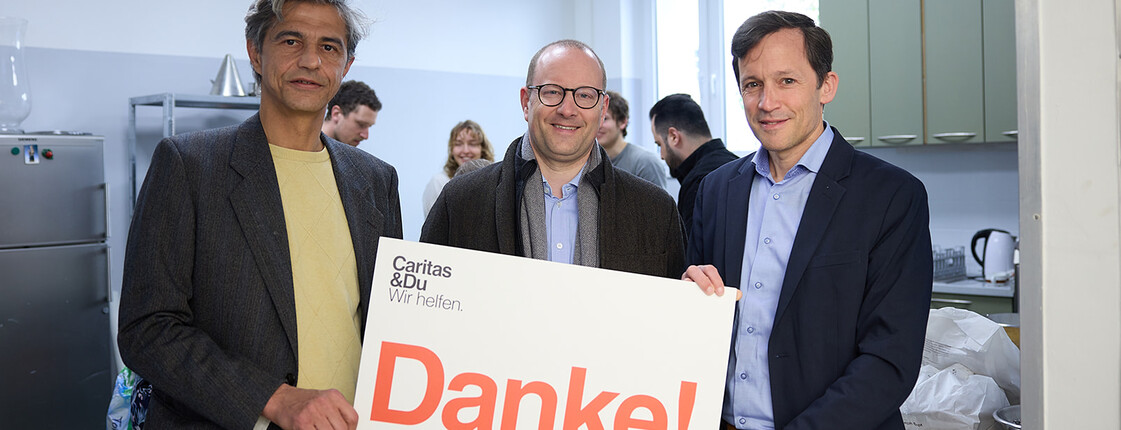 Drei Personen stehen in einem Küchenraum und halten gemeinsam ein Schild mit der Aufschrift „Danke!“ von Caritas & Du – Wir helfen.