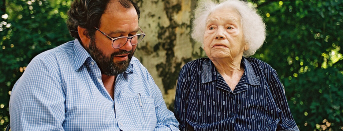 Ein Mann mittleren Alters sitzt neben einer alten Dame. Die Dame trägt eine blaue Kleidung und sitzt im Rollstuhl. Der Herr liest ihr etwas aus einem dicken Buch vor und die Dame hört ihm gespannt zu.