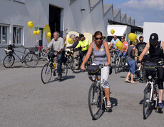 Ca. 20 Menschen mit an ihren Rädern gelb befestigten Luftballonen befinden sich auf einem großen asphaltierten Platz. 