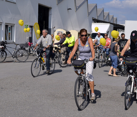 Ca. 20 Menschen mit an ihren Rädern gelb befestigten Luftballonen befinden sich auf einem großen asphaltierten Platz. 