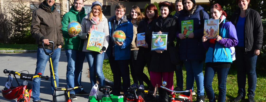 Eine Gruppe junger Erwachsener steht draußen vor einem Wohnblock und zeigt stolz Spielzeug, das die Kinder der MUKI bekommen. Darunter befinden sich Spielzeugtraktoren und Fahrräder mit Stützrädern. Auch Bücher sind dabei. 
