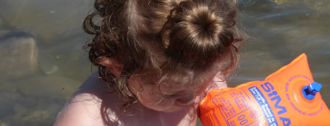Ein kleines Kind mit orangen Schwimmflügeln kühlt sich im seichten Wasser ab. Die braunen Haare hat es mit einem Dutt nach oben zusammengebunden.  