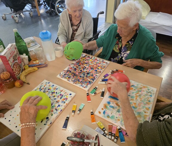 Mehrere Bewohnerinnen des Pflegewohnhauses experimentieren kreativ mit Farben und Luftballons, um lebendige und farbenfrohe Kunstwerke auf Leinwänden zu gestalten.