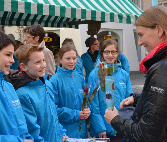 Eine junge Frau steht neben jungen Kindern und hält rote Rosen in der Hand. Die Kinder strahlen.