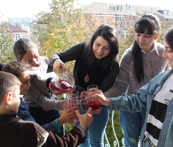 Eine Betreuerin schenkt einer Gruppe Kinder und Jugendlicher roten Saft aus einer Glaskaraffe aus.