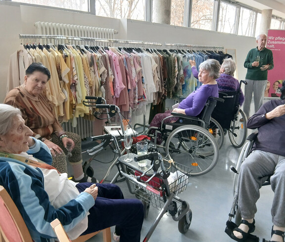 Gruppe von Bewohner*innen sitzt vor Kleiderständern mit farbiger Mode