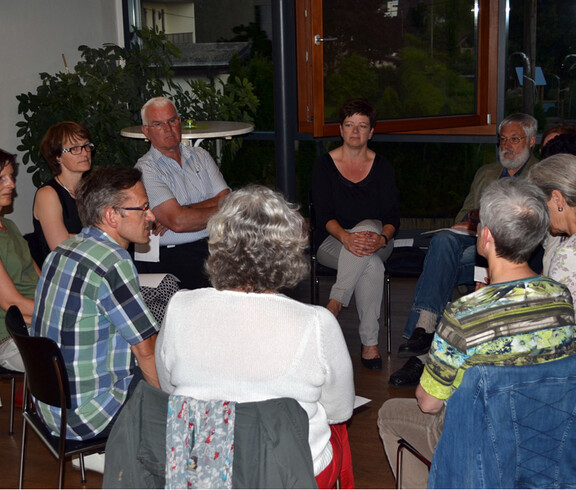 Eine Gruppe von etwa zehen Personen sind in einem Sitzkreis zusammengekommen. Dabei sind es überwiegend ältere Mitmenschen. 