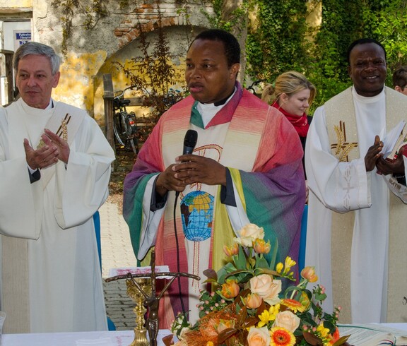 Drei Pfarrer stehen hinter einem Tisch, der mittlere trägt bunte Kleidung während die beiden anderen in Weiß gekleidet sind.