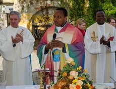 Drei Pfarrer stehen hinter einem Tisch, der mittlere trägt bunte Kleidung während die beiden anderen in Weiß gekleidet sind.