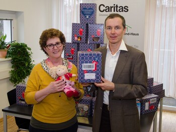 Zwei Personen halten eine Schachtel und eine Puppe in die Kamera. Hinter ihnen sind noch mehrere Schachteln.