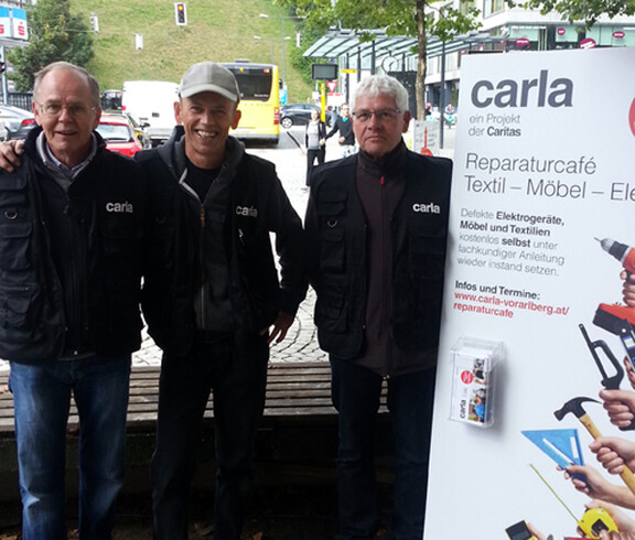 Drei carla Mitarbeiter vom Reparaturcafe posieren zu einem gemeinsamen Gruppenfoto in der feldkicher Innenstadt. Neben ihnen steht ein weißes carla Roll-Up. Die Herren mittleren Alters sind größtenteils schwarz gekleidet. Auf ihren schwarzen Weste steht in Weiß carla. 