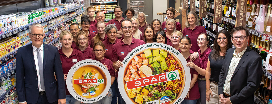 Große Gruppe in Supermarkt hält runde Tafeln mit SPAR-Logo und Karitas-Hinweis im Gang.